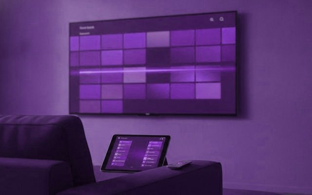 A TV showing a programme guide grid with a tablet and phone showing channel lists on a sofa