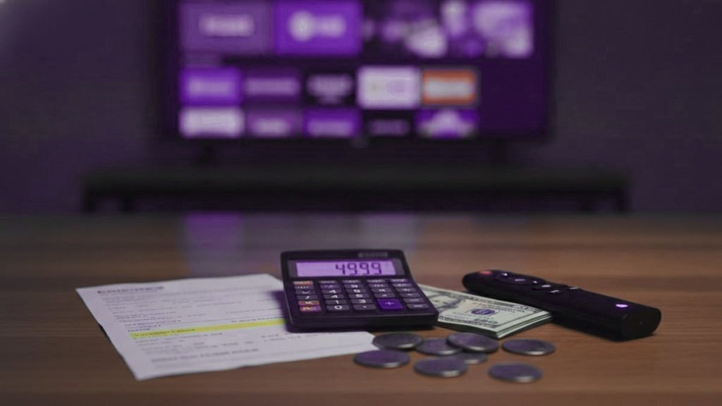 A calculator, dollar bills, coins, a streaming stick and a remote on a table in front of a TV