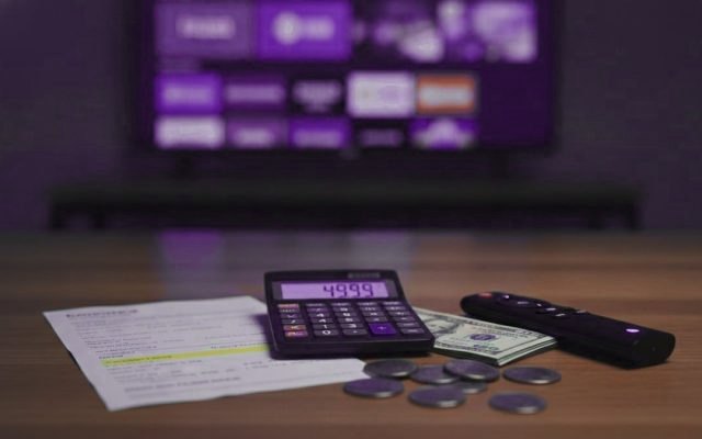 A calculator, dollar bills, coins, a streaming stick and a remote on a table in front of a TV